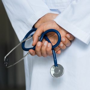 A doctor wearing a white coat and holding a stethoscope behind their back