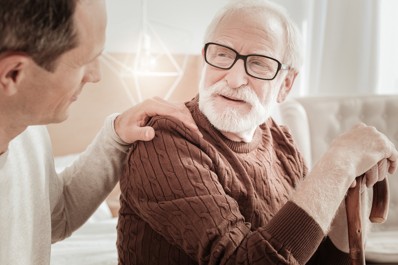 Aged man sitting on the bed supporting on the cane and listening to his son.