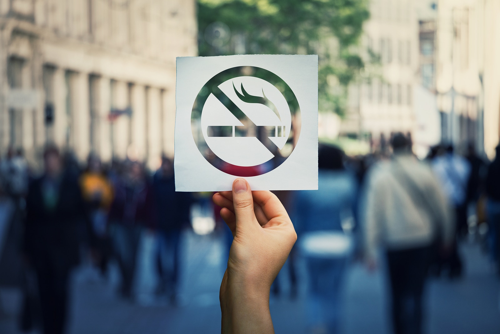 Hand Holding A Paper Sheet With No Smoking Sign Over A Crowded Street